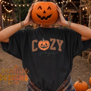 May include: A black short-sleeve t-shirt featuring a graphic print with the text "COZY season" in a checkered orange and black pattern. The letter "O" is replaced by an orange jack-o'-lantern illustration with a carved face and stem.
