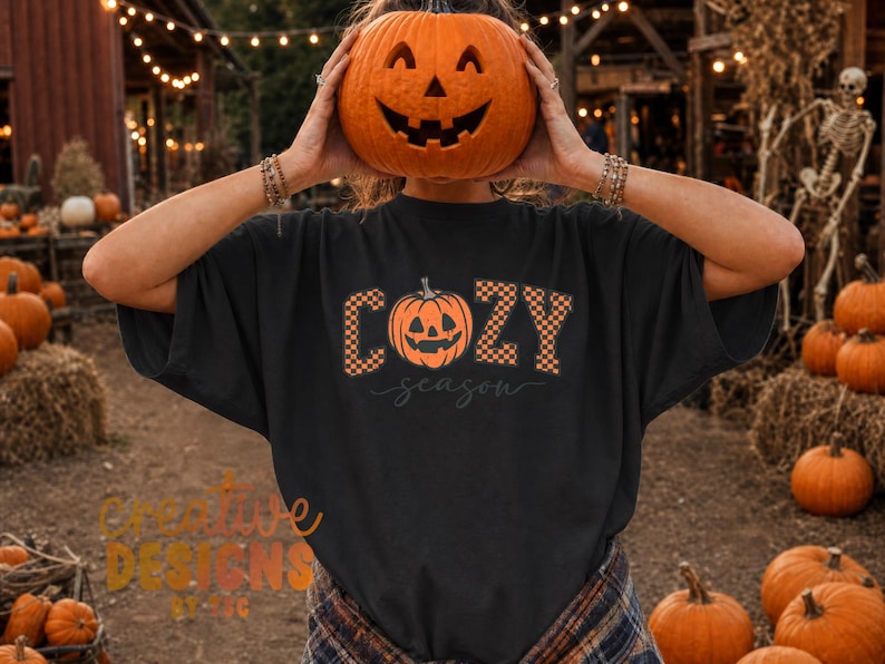 May include: A black short-sleeve t-shirt featuring a graphic print with the text "COZY season" in a checkered orange and black pattern. The letter "O" is replaced by an orange jack-o'-lantern illustration with a carved face and stem.