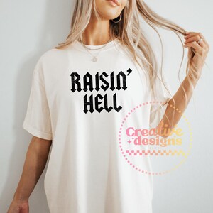 May include: A cream-coloured t-shirt featuring the text "Raisin' Hell" in black gothic font. The shirt has a loose fit and short sleeves. A circular logo with text is visible on the lower right.