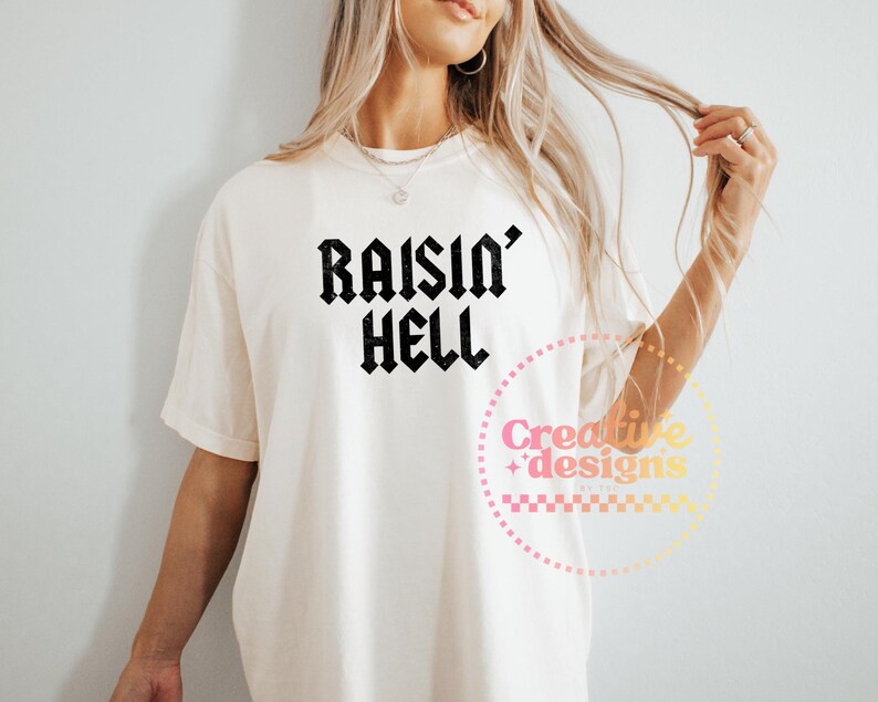 May include: A cream-coloured t-shirt featuring the text "Raisin' Hell" in black gothic font. The shirt has a loose fit and short sleeves. A circular logo with text is visible on the lower right.