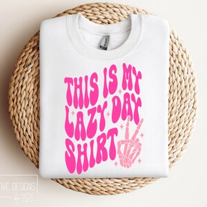 Puede incluir: Sudadera blanca con texto rosa que dice "This is my lazy day shirt" con una mano de esqueleto rosa haciendo un signo de paz.
