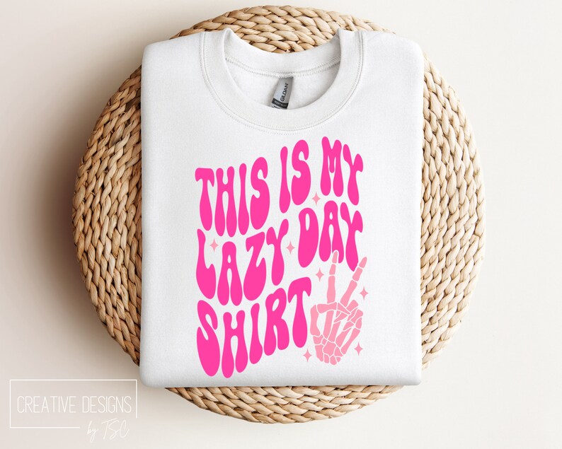 Puede incluir: Sudadera blanca con texto rosa que dice "This is my lazy day shirt" con una mano de esqueleto rosa haciendo un signo de paz.