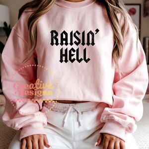 May include: A pale pink sweatshirt featuring the text "RAISIN' HELL" in a black gothic font. The sweatshirt has a crew neck and long sleeves. The model is wearing white sweatpants.