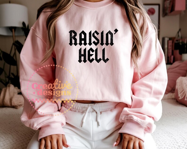May include: A pale pink sweatshirt featuring the text "RAISIN' HELL" in a black gothic font. The sweatshirt has a crew neck and long sleeves. The model is wearing white sweatpants.