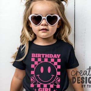 May include: A black short-sleeved t-shirt featuring a pink graphic print of a checkered square containing a smiley face making a peace sign. The text "BIRTHDAY GIRL" is printed in pink above the graphic. The shirt is worn with white heart-shaped sunglasses.