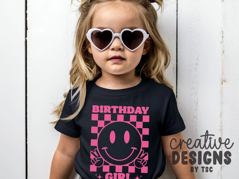 May include: A black short-sleeved t-shirt featuring a pink graphic print of a checkered square containing a smiley face making a peace sign. The text "BIRTHDAY GIRL" is printed in pink above the graphic. The shirt is worn with white heart-shaped sunglasses.