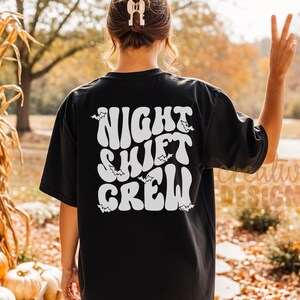 May include: A black short-sleeved t-shirt featuring white text on the back that reads "NIGHT SHIFT CREW" in a wavy, retro-inspired font. Small white bat silhouettes are scattered around the lettering, which is centered on the back of the shirt.