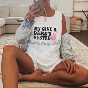 May include: White tank top with the text "My Give a Damn's Busted" in black lettering and a pink peace sign.