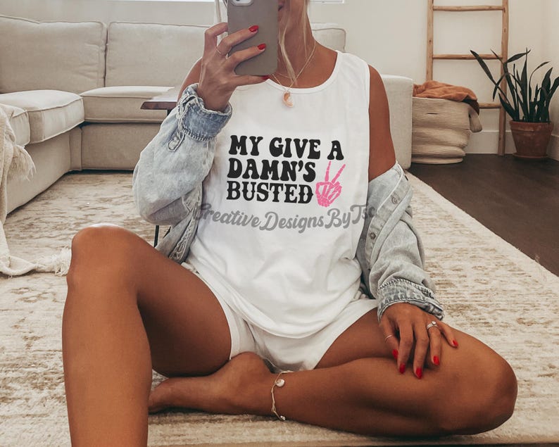 May include: White tank top with the text "My Give a Damn's Busted" in black lettering and a pink peace sign.