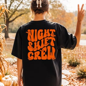 May include: A black short-sleeved t-shirt featuring the orange text NIGHT SHIFT CREW in a wavy, retro-inspired font on the back. The design includes small orange bat silhouettes scattered among the letters of the text.