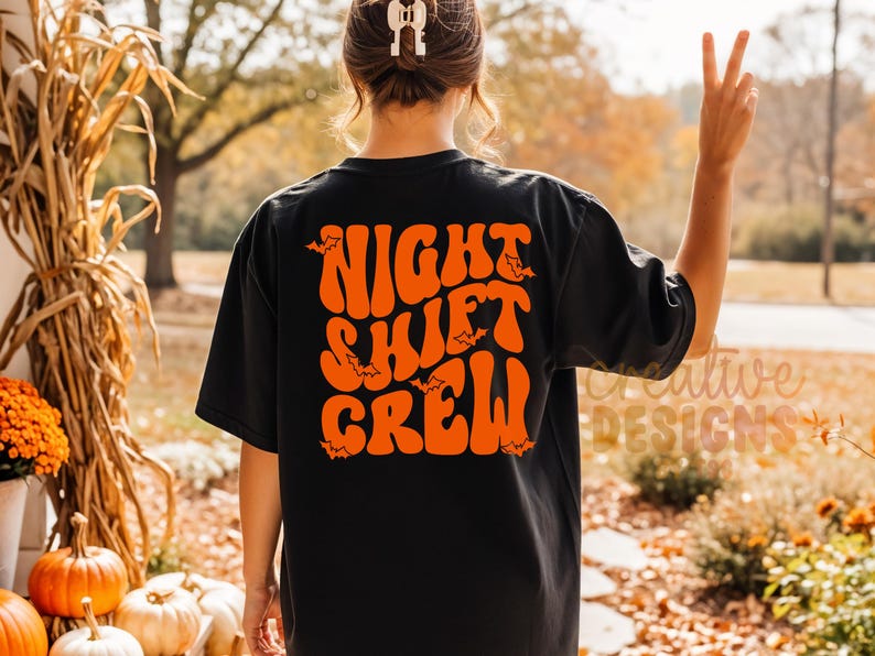 May include: A black short-sleeved t-shirt featuring the orange text NIGHT SHIFT CREW in a wavy, retro-inspired font on the back. The design includes small orange bat silhouettes scattered among the letters of the text.