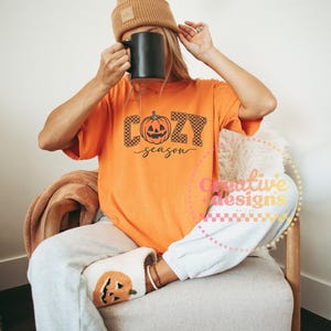 May include: Orange t-shirt with the words "Cozy Season" and a pumpkin graphic. The person is wearing a brown beanie, gray sweatpants, and pumpkin-themed slippers. A black mug is held up.