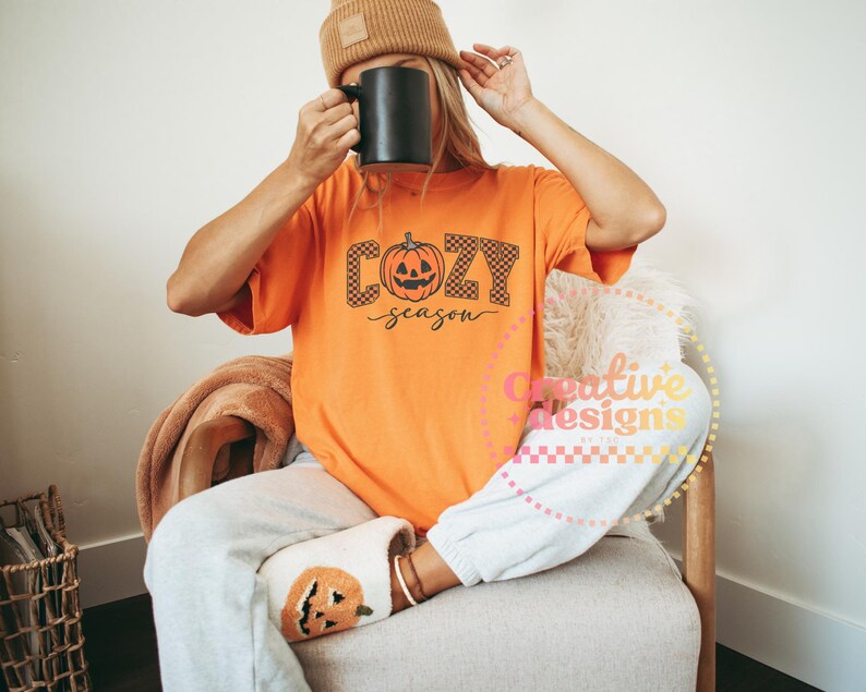 May include: Orange t-shirt with the words "Cozy Season" and a pumpkin graphic. The person is wearing a brown beanie, gray sweatpants, and pumpkin-themed slippers. A black mug is held up.