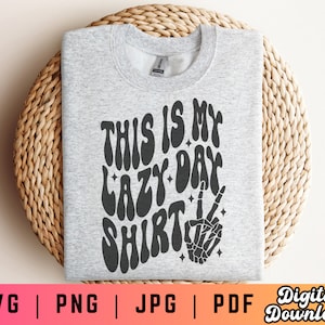 Puede incluir: Una sudadera gris con el texto "This is my lazy day shirt" en una fuente retro. Una mano de esqueleto negro con un signo de paz est&aacute; debajo del texto.
