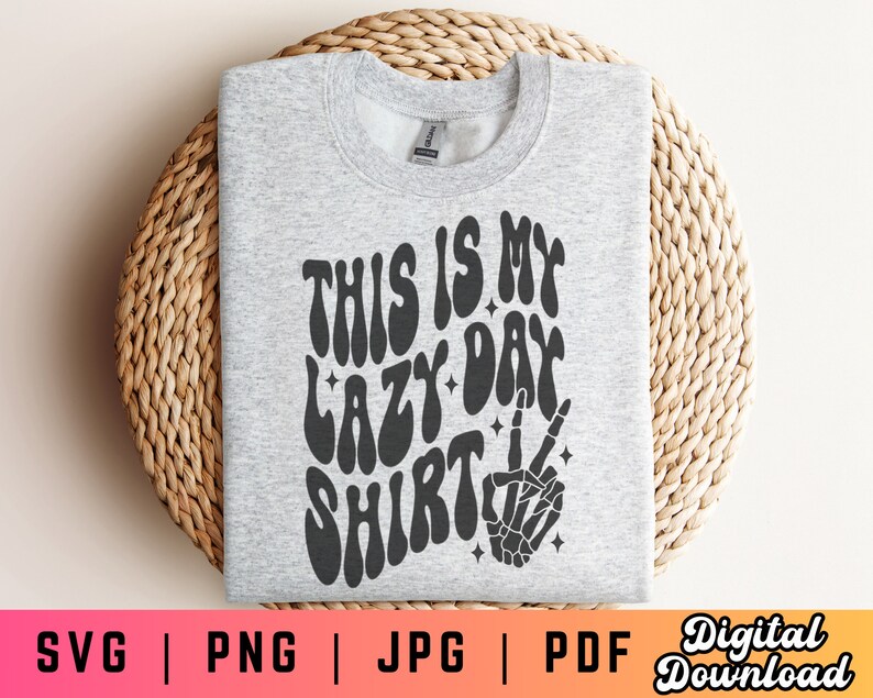 Puede incluir: Una sudadera gris con el texto "This is my lazy day shirt" en una fuente retro. Una mano de esqueleto negro con un signo de paz est&aacute; debajo del texto.