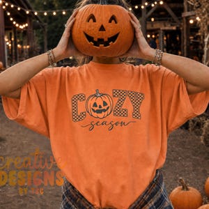 May include: An orange t-shirt featuring a black graphic print of a jack-o'-lantern pumpkin centered between the words COZY and season. The letters COZY are filled with a black and white checkerboard pattern, while the word season is in script.