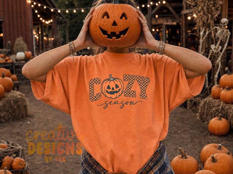 May include: An orange t-shirt featuring a black graphic print of a jack-o'-lantern pumpkin centered between the words COZY and season. The letters COZY are filled with a black and white checkerboard pattern, while the word season is in script.