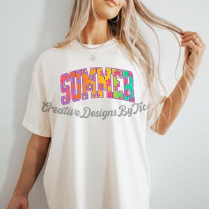 Puede incluir: Una camiseta blanca con la palabra "SUMMER" impresa en letras de colores con un patr&oacute;n floral. Las letras est&aacute;n delineadas en morado y tienen un estilo retro.
