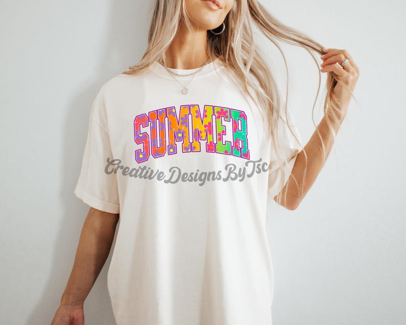 Puede incluir: Una camiseta blanca con la palabra "SUMMER" impresa en letras de colores con un patr&oacute;n floral. Las letras est&aacute;n delineadas en morado y tienen un estilo retro.