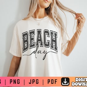 May include: A white t-shirt with the text "Beach Day" in black lettering. The text is in a bold, blocky font with a slight outline.