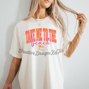 K&ouml;nnte beinhalten: Wei&szlig;es T-Shirt mit dem Text "Take Me To The Beach" in orangefarbener und rosafarbener Schrift. Unter dem Text befindet sich eine kleine Palme und ein Sonnenmotiv.