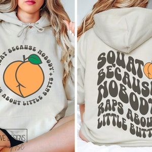 K&ouml;nnte beinhalten: Ein hellbeiger Hoodie mit einer Pfirsichgrafik und dem Text "Squat because nobody raps about little butts" in einer Retro-Schrift.