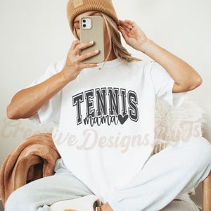 May include: White t-shirt with black text that says "Tennis Mama" with a black heart. The model is wearing a brown beanie and holding a phone in front of their face.