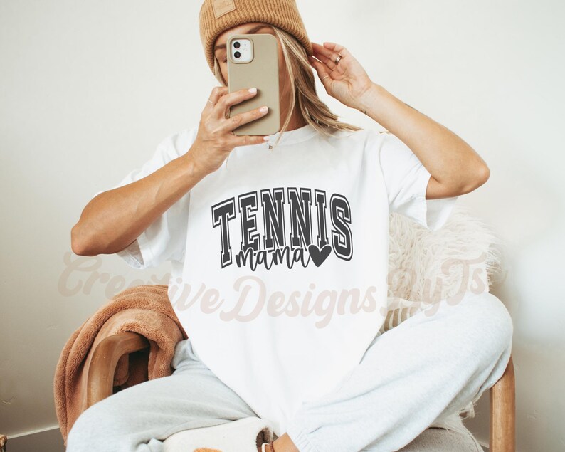 May include: White t-shirt with black text that says "Tennis Mama" with a black heart. The model is wearing a brown beanie and holding a phone in front of their face.