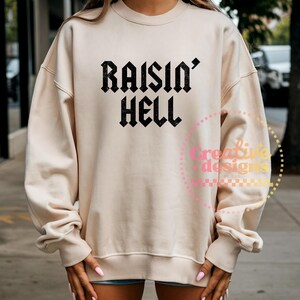 May include: A beige sweatshirt featuring the text "RAISIN' HELL" in black, gothic font. The long-sleeved top has a crew neck and ribbed cuffs. The sweatshirt is a casual style.