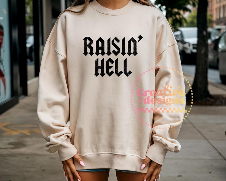 May include: A beige sweatshirt featuring the text "RAISIN' HELL" in black, gothic font. The long-sleeved top has a crew neck and ribbed cuffs. The sweatshirt is a casual style.