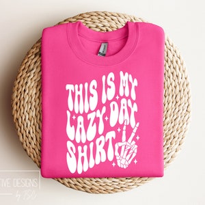 Puede incluir: Sudadera rosa con texto blanco que dice "This is my lazy day shirt" en una fuente retro. Una mano de esqueleto blanca con un signo de paz est&aacute; a la derecha del texto.