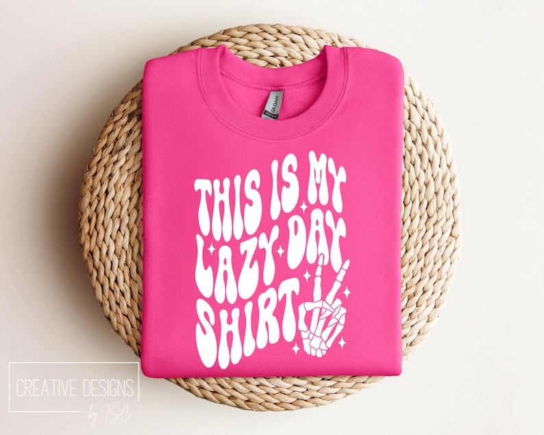 Puede incluir: Sudadera rosa con texto blanco que dice "This is my lazy day shirt" en una fuente retro. Una mano de esqueleto blanca con un signo de paz est&aacute; a la derecha del texto.