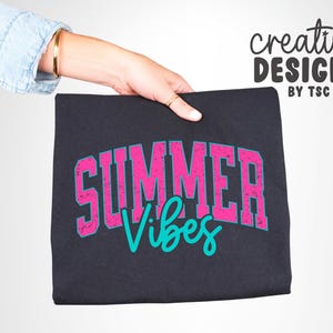 May include: A black t-shirt with the words "SUMMER Vibes" in a distressed pink font with a teal outline. The words are in a collegiate style font. The words "creative DESIGNS BY TSC" are in black script in the upper right corner.