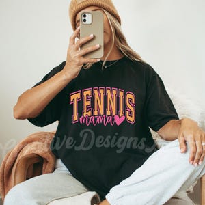 May include: A black t-shirt with the text "Tennis Mama" in orange and pink lettering with a pink heart. The shirt is being worn by a person with a brown beanie.