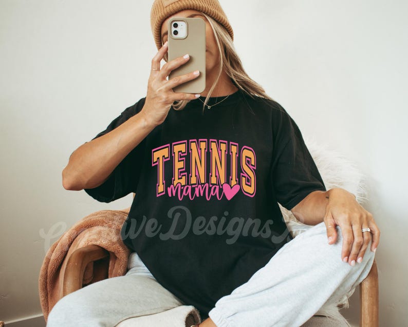 May include: A black t-shirt with the text "Tennis Mama" in orange and pink lettering with a pink heart. The shirt is being worn by a person with a brown beanie.