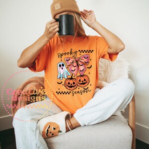 Puede incluir: Camiseta naranja con un dise&ntilde;o de Halloween. El dise&ntilde;o incluye un fantasma, un lazo, dos calabazas y la frase "spooky season". La camiseta se lleva con un pantal&oacute;n de ch&aacute;ndal gris y un gorro marr&oacute;n.