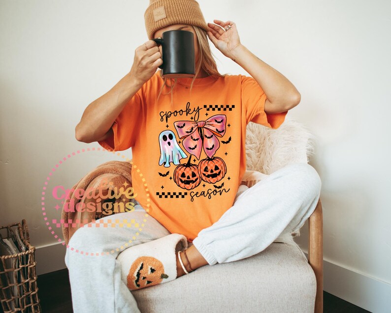 Puede incluir: Camiseta naranja con un dise&ntilde;o de Halloween. El dise&ntilde;o incluye un fantasma, un lazo, dos calabazas y la frase "spooky season". La camiseta se lleva con un pantal&oacute;n de ch&aacute;ndal gris y un gorro marr&oacute;n.