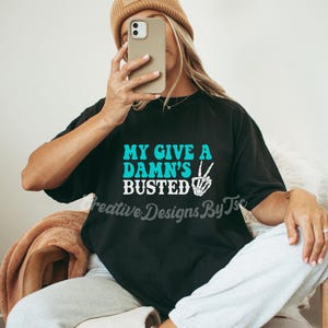 May include: Black t-shirt with the text "My Give A Damn's Busted" in turquoise blue letters with a white peace sign and skeleton hand graphic.