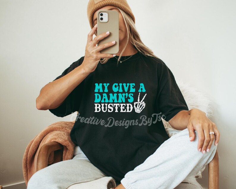 May include: Black t-shirt with the text "My Give A Damn's Busted" in turquoise blue letters with a white peace sign and skeleton hand graphic.