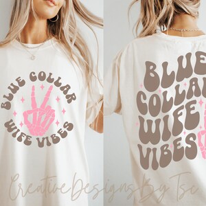 May include: A white t-shirt with the text "BLUE COLLAR WIFE VIBES" in brown and pink lettering with a pink skeleton hand making a peace sign in the center of the circle. The text is in a retro font style.