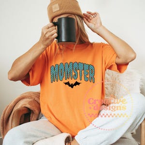 May include: Orange t-shirt with the word "MONSTER" in blue and green bubble letters and a black bat graphic. The shirt is part of a Halloween-themed collection.