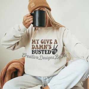 May include: A white sweatshirt with the text "My Give A Damn's Busted" in brown and black lettering. A peace sign with a skeleton hand is next to the text.
