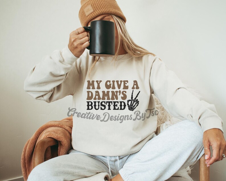 May include: A white sweatshirt with the text "My Give A Damn's Busted" in brown and black lettering. A peace sign with a skeleton hand is next to the text.