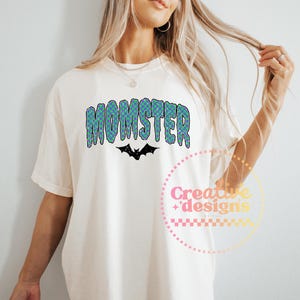 May include: Cream-colored t-shirt with the word "MOMSTER" in a blue and green checkered pattern, with a black bat graphic underneath. The shirt is a casual style.