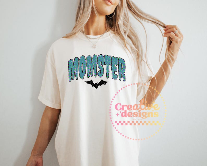 May include: Cream-colored t-shirt with the word "MOMSTER" in a blue and green checkered pattern, with a black bat graphic underneath. The shirt is a casual style.
