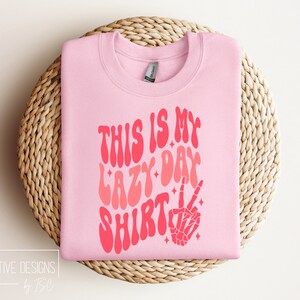 Puede incluir: Sudadera rosa con el texto "This is my lazy day shirt" en una fuente retro con un efecto degradado rosa y rojo. Una mano de esqueleto rosa con un signo de paz est&aacute; a la derecha del texto.