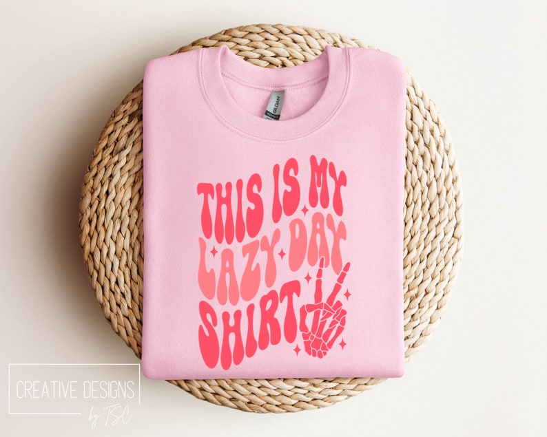Puede incluir: Sudadera rosa con el texto "This is my lazy day shirt" en una fuente retro con un efecto degradado rosa y rojo. Una mano de esqueleto rosa con un signo de paz est&aacute; a la derecha del texto.