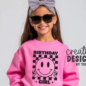May include: A bright pink sweatshirt featuring a black and pink checkerboard square graphic with a central smiley face making peace signs. The text "BIRTHDAY GIRL" is printed in black above and below the graphic, accompanied by small star icons.