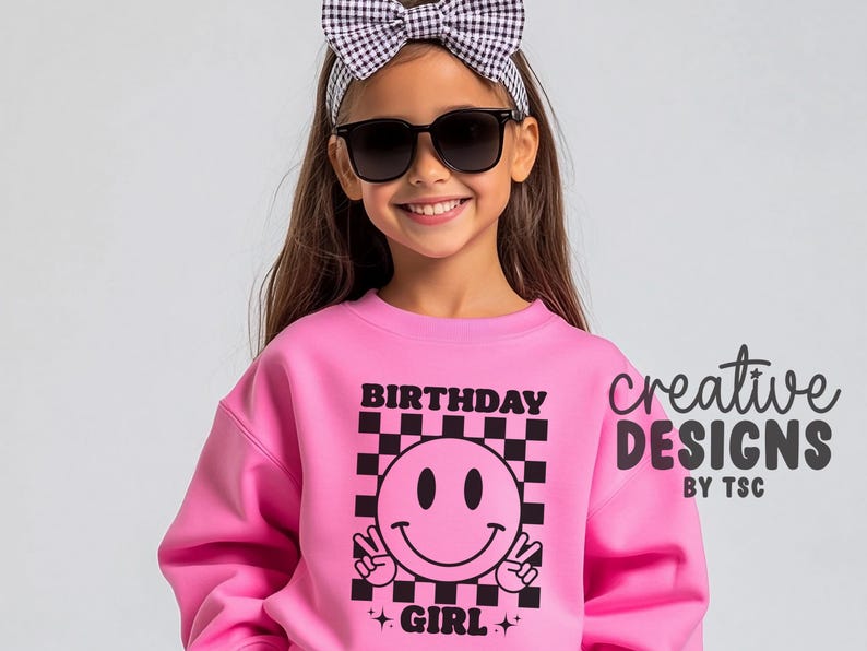 May include: A bright pink sweatshirt featuring a black and pink checkerboard square graphic with a central smiley face making peace signs. The text "BIRTHDAY GIRL" is printed in black above and below the graphic, accompanied by small star icons.