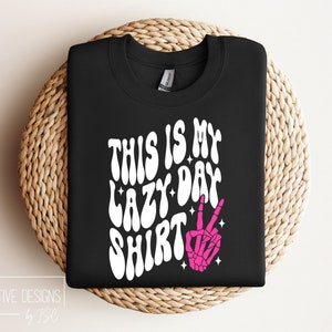 Puede incluir: Camiseta negra con texto blanco que dice "This is my lazy day shirt" en una fuente retro. Una mano de esqueleto rosa con un signo de paz est&aacute; a la derecha del texto.